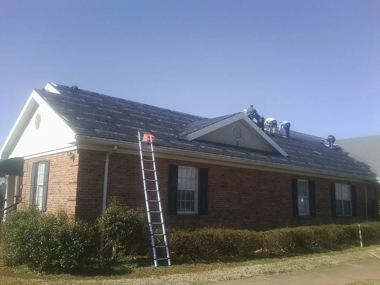 Crew tearing shingles off a brick ranch roof with a ladder set — Metal Roof Repair work by our crew in Wellford