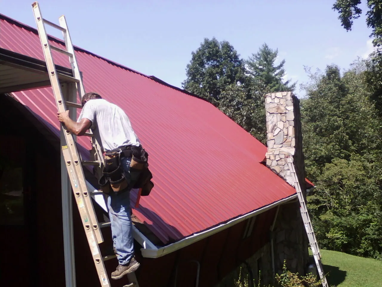 Roofer on a ladder beside a red metal roof and stone chimney — Soffit & Fascia Repair by Roofing Brothers in Wellford